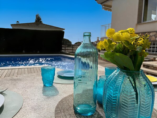 Une table avec une bouteille, des verres et un vase rempli de fleurs, près d’une piscine.