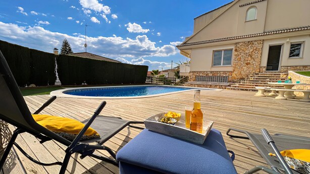 Un área junto a la piscina con sillas de descanso, una mesa con bebidas y un edificio de piedra al fondo.