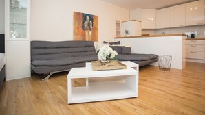 Una sala de estar moderna con un sofá gris, una mesa de centro blanca y una zona de cocina con armarios blancos.