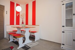 Un espace repas avec une table et des bancs blancs, des coussins rouges et un vase rempli de fleurs.