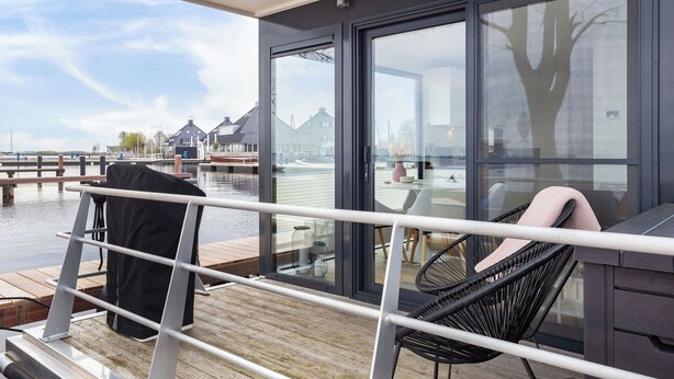 Une terrasse dotée d’une porte vitrée donnant sur une table et des chaises, avec vue sur une étendue d’eau et des maisons en arrière-plan.
