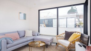 Un salon moderne avec un canapé gris, un fauteuil en osier, une petite table ronde et une grande fenêtre donnant sur la ville.