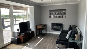 A modern living room with a flat-screen TV, a sofa, a fireplace, and a cityscape painting. - Cosy, self-contained rural bungalow – peaceful countryside escape (Out Rawcliffe Preston)