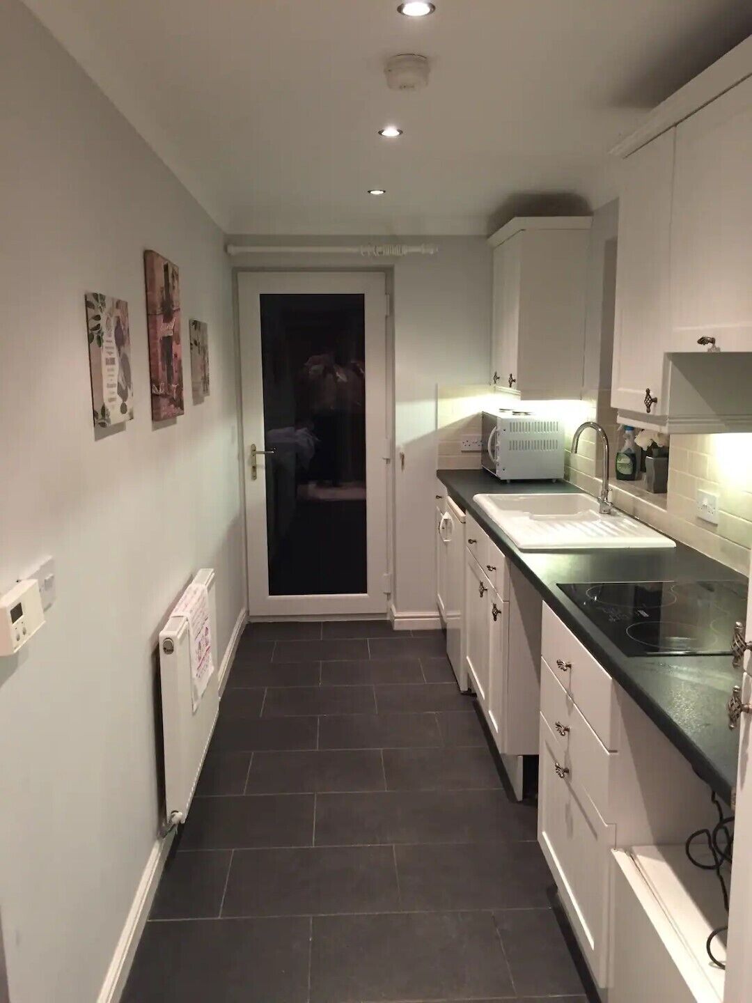 A narrow kitchen with white cabinets, a black countertop, and a door leading to a dark room.