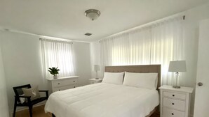 A spacious bedroom with a large bed, two bedside tables with lamps, a bench, and a window with sheer curtains.