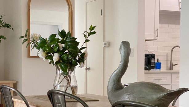 A dining area with a wooden table, metal chairs, a vase with flowers, and a kitchen in the background.