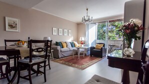 Een woonkamer met een bank, stoelen, een eettafel met een fruitschaal, een kroonluchter en een raam met uitzicht op bomen.