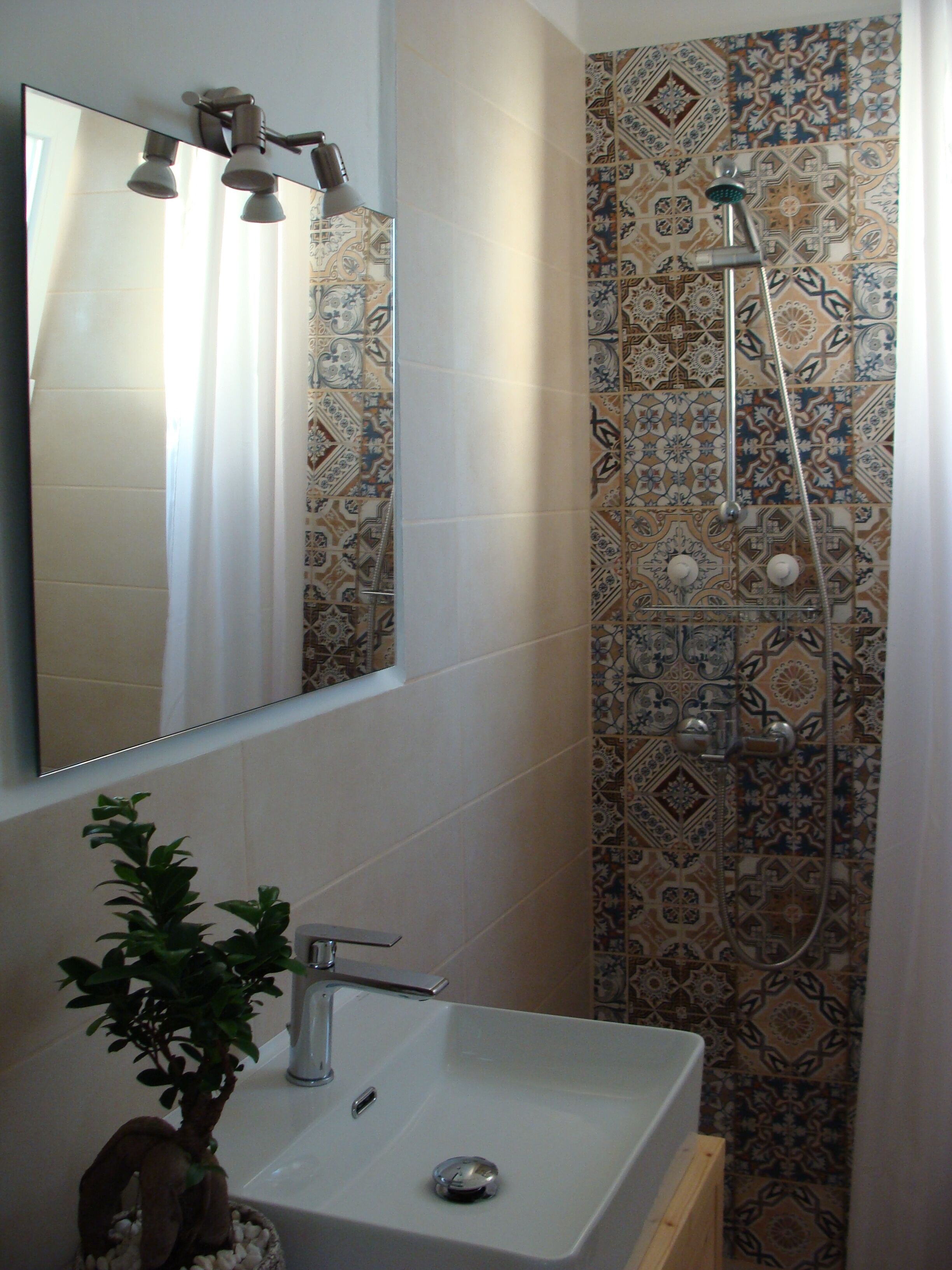 Een badkamer met een witte wastafel, een spiegel, een douche met een handdouche en betegelde muren.