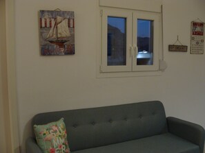 Un salon avec un canapé gris, un tableau représentant un voilier, une fenêtre à deux vitres et un panneau indiquant «
Il est interdit de fumer
».