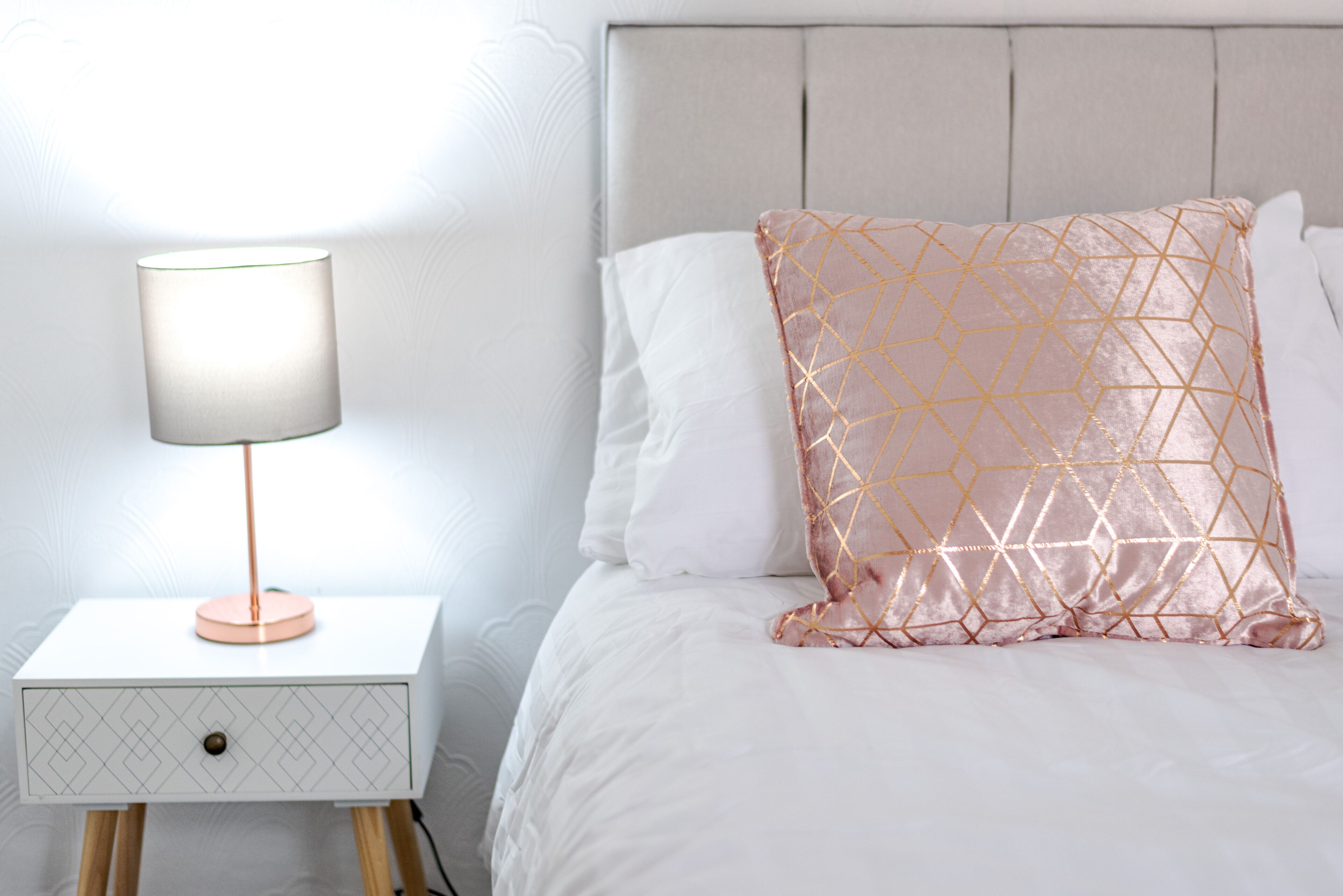 Una cama bien hecha con cabecera blanca, lámpara de noche y almohada decorativa.