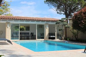 A swimming pool with a tiled deck, a covered patio area with chairs, and a large tree in the background.