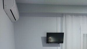 A small, simple room with a bed, a sofa, a TV mounted on the wall, and an air conditioning unit. - Ilio Socrates Triple room with sea view. (Thassos)