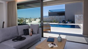 Uma sala de estar moderna com um sofá, uma mesa de centro com um laptop, óculos de sol e um livro. Uma grande janela oferece vista para uma piscina e uma parede de pedra.