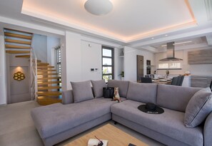 Un salon moderne avec un canapé d’angle gris, un escalier en bois et une cuisine en arrière-plan.