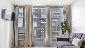 Ein modernes Wohnzimmer mit grauem Sofa, einem Flachbildfernseher an der Wand und einem großen Fenster mit Vorhängen.