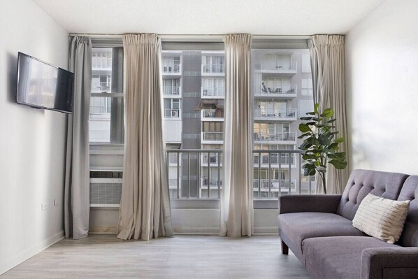 A modern living room with a grey sofa, a flat-screen TV mounted on the wall, and a large window with curtains.