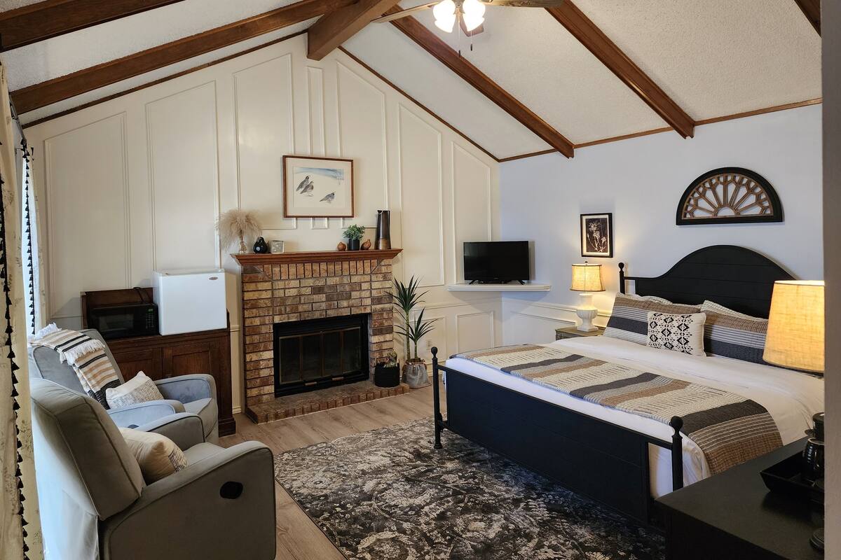 A bedroom with a fireplace, a bed with a headboard, a television, and a ceiling fan.