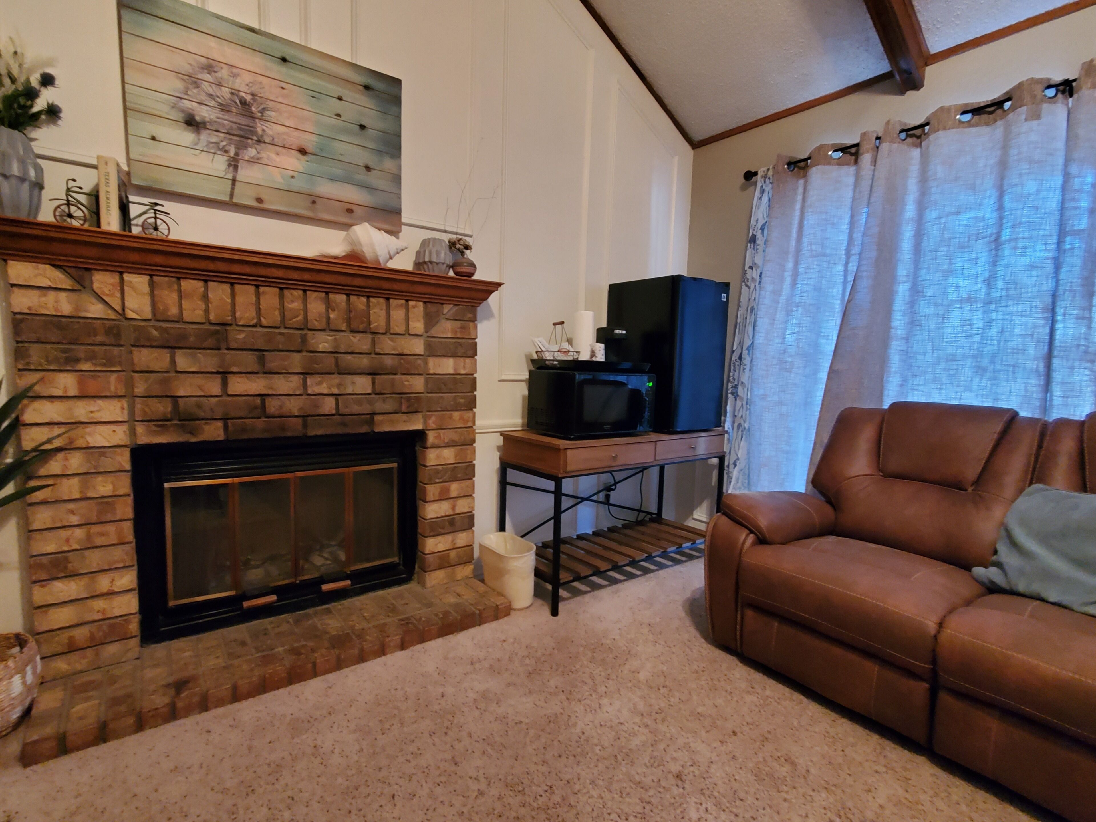 Una sala de estar con chimenea de ladrillo, un sofá de cuero marrón, un televisor sobre un mueble de madera y una ventana con cortinas.