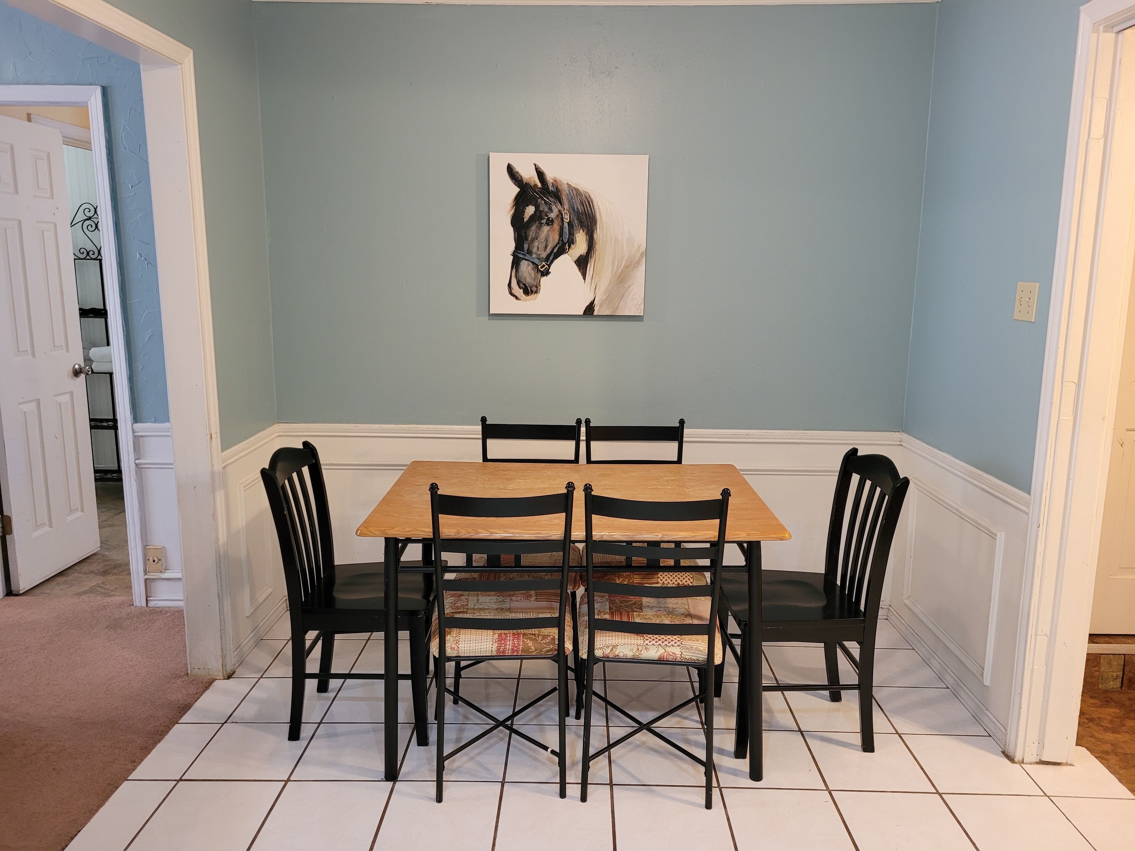 Un comedor con una mesa y sillas, un cuadro de un caballo en la pared, y una puerta que da a otra habitación.