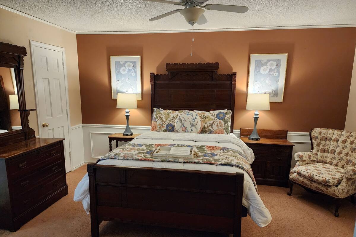 A bedroom with a wooden bed, two bedside lamps, a chair, and floral artwork on the walls.
