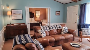 A living room with a brown leather sofa, a wooden side table, and a chair. There is a door leading to a bedroom with a bed and a chair. - Butterfield Inn Main Street Cottages (Fort Davis)
