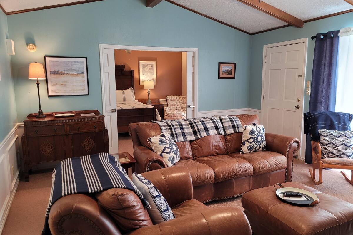 A living room with a brown leather sofa, a wooden side table, and a chair. There is a door leading to a bedroom with a bed and a chair.