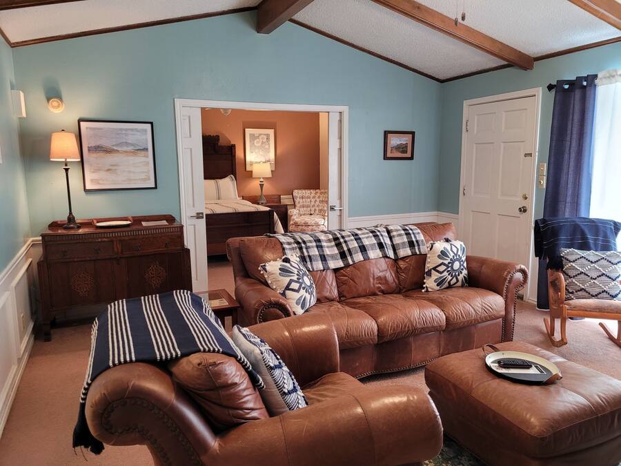 Sala de estar com um sofá de couro marrom, uma mesinha de madeira e uma cadeira. Há uma porta que dá para um quarto com uma cama e uma cadeira.