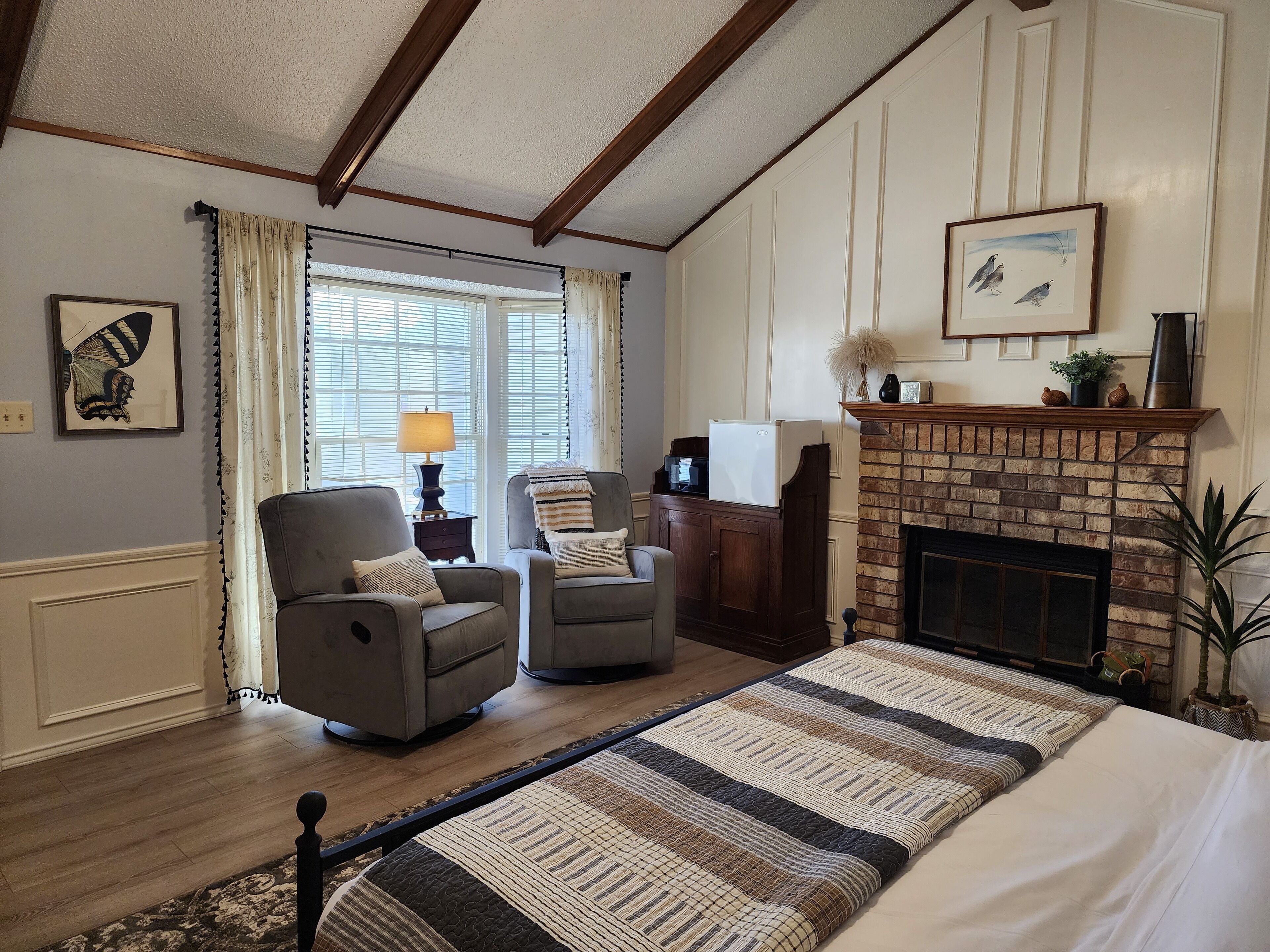 A cozy living room with a fireplace, two armchairs, a large bed, and a window with curtains.