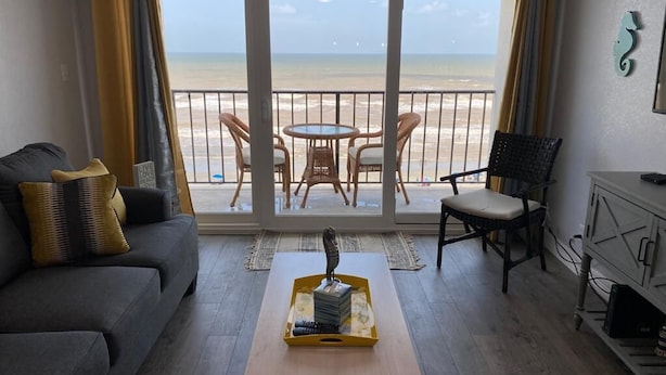 A living room with a sofa, a coffee table with a decorative item, a dining table with chairs, and a view of the beach through large windows. - Oceanview Beach Getaway, Great balcony!! (Galveston)