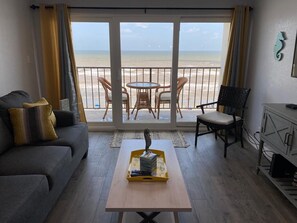 Ett vardagsrum med en soffa, ett soffbord med en dekorativ detalj, ett matbord med stolar och utsikt över stranden genom stora fönster.