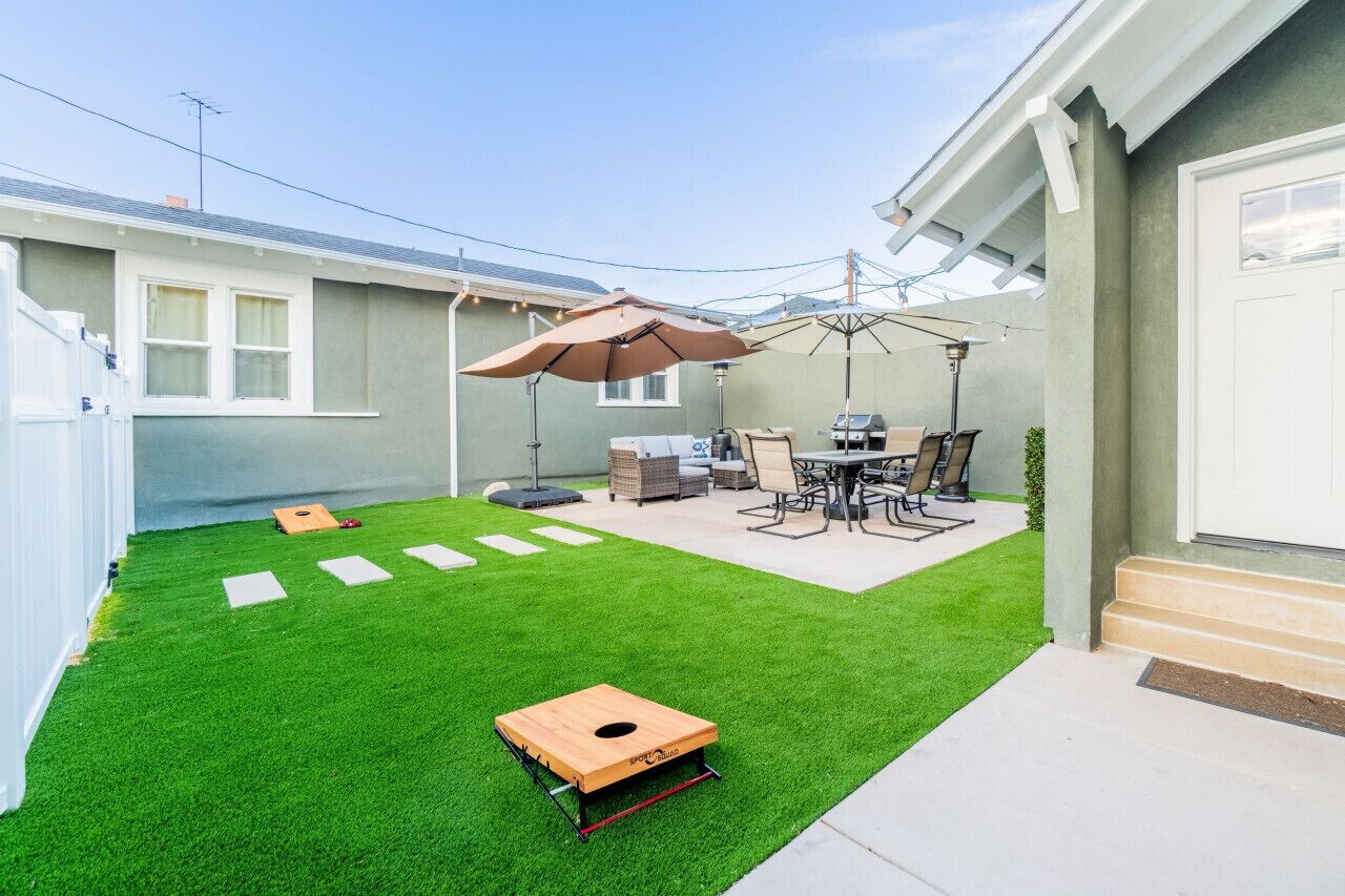 Un patio con césped artificial, una estructura de juego de madera, una parrilla y mobiliario exterior.