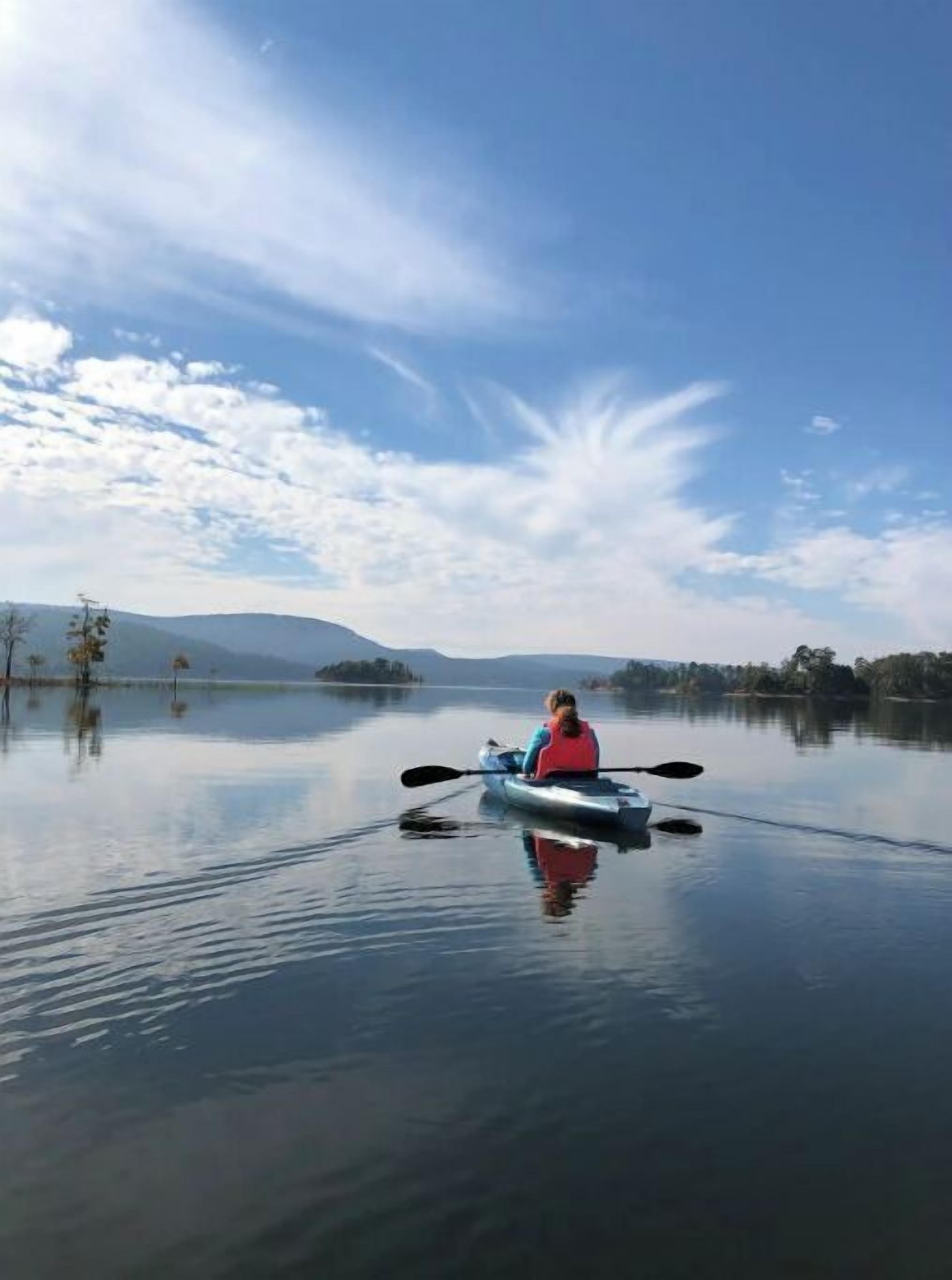 Eine Person paddelt mit einem Kajak auf einem ruhigen See, im Hintergrund sind Berge zu sehen.