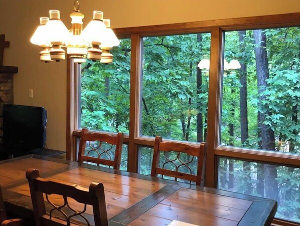 Enjoy meals in this dining area with a large table and four chairs, overlooking a dense forest through a large window.