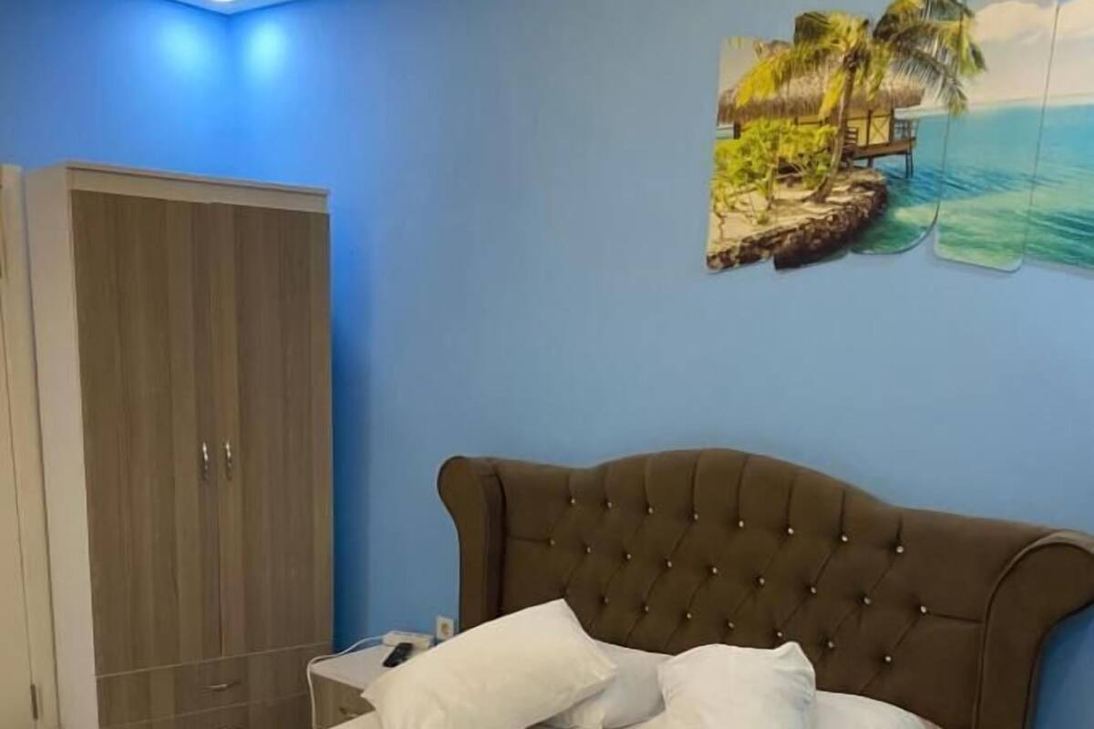A neatly made bed with white linens and pillows, a wooden headboard, and a tropical mural on the wall.