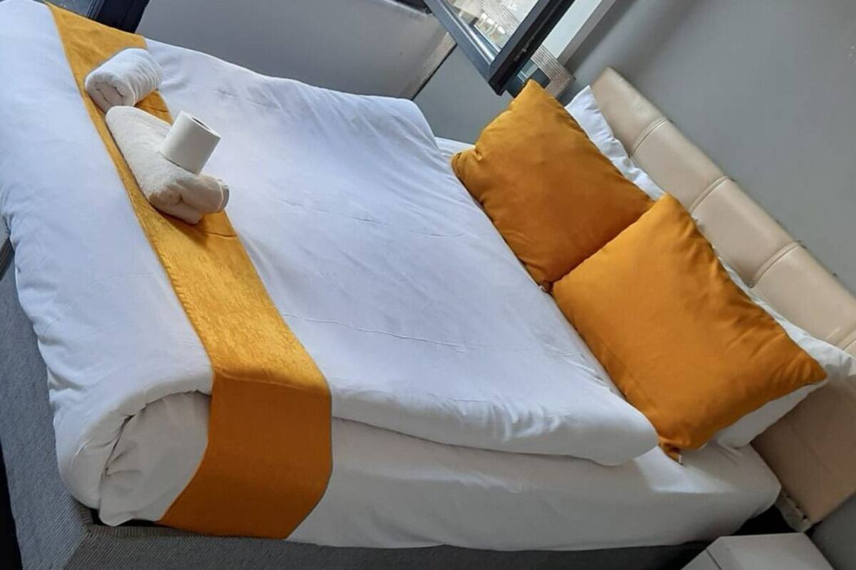 A neatly made bed with white and yellow bedding, a rolled towel, and two orange pillows.