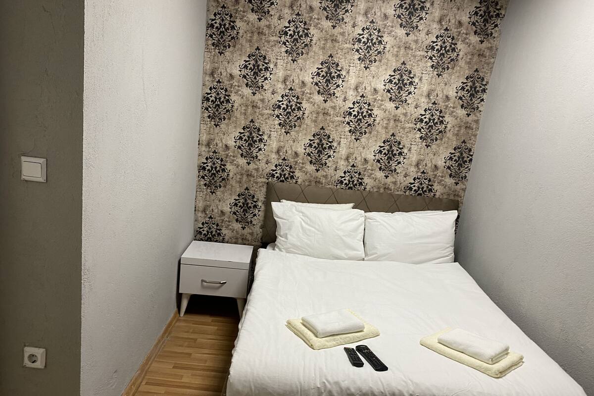 A neatly made bed with white linens and two folded towels, a bedside table with a lamp, and a patterned wallpaper on the wall.