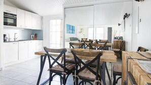 Une cuisine moderne avec des meubles blancs, un four encastré et une machine à café. On y trouve des tables et des chaises en bois, dotées de sièges rembourrés.