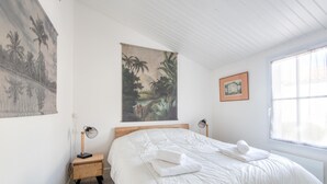 Une chambre avec un grand lit, deux tables de chevet, un ventilateur fixé au mur, une fenêtre avec des rideaux légers et un tableau sur le thème des tropiques.