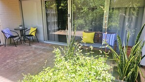 Eine Terrasse mit Backsteinboden, eine Glastür, die in einen Garten führt, und ein Sitzbereich mit einer Bank und Stühlen.