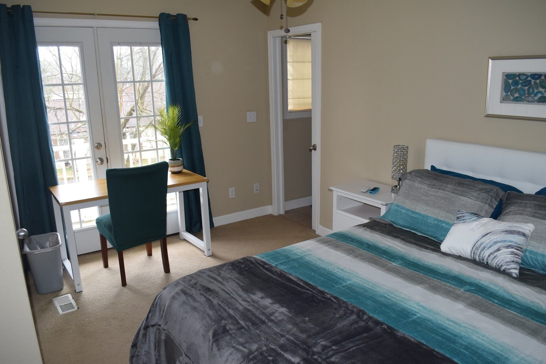 A bedroom with a bed, a desk, and a chair. There is a window with curtains, a door, and a wall-mounted lamp.