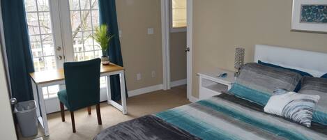 A bedroom with a bed, a desk, and a chair. There is a window with curtains, a door, and a wall-mounted lamp.