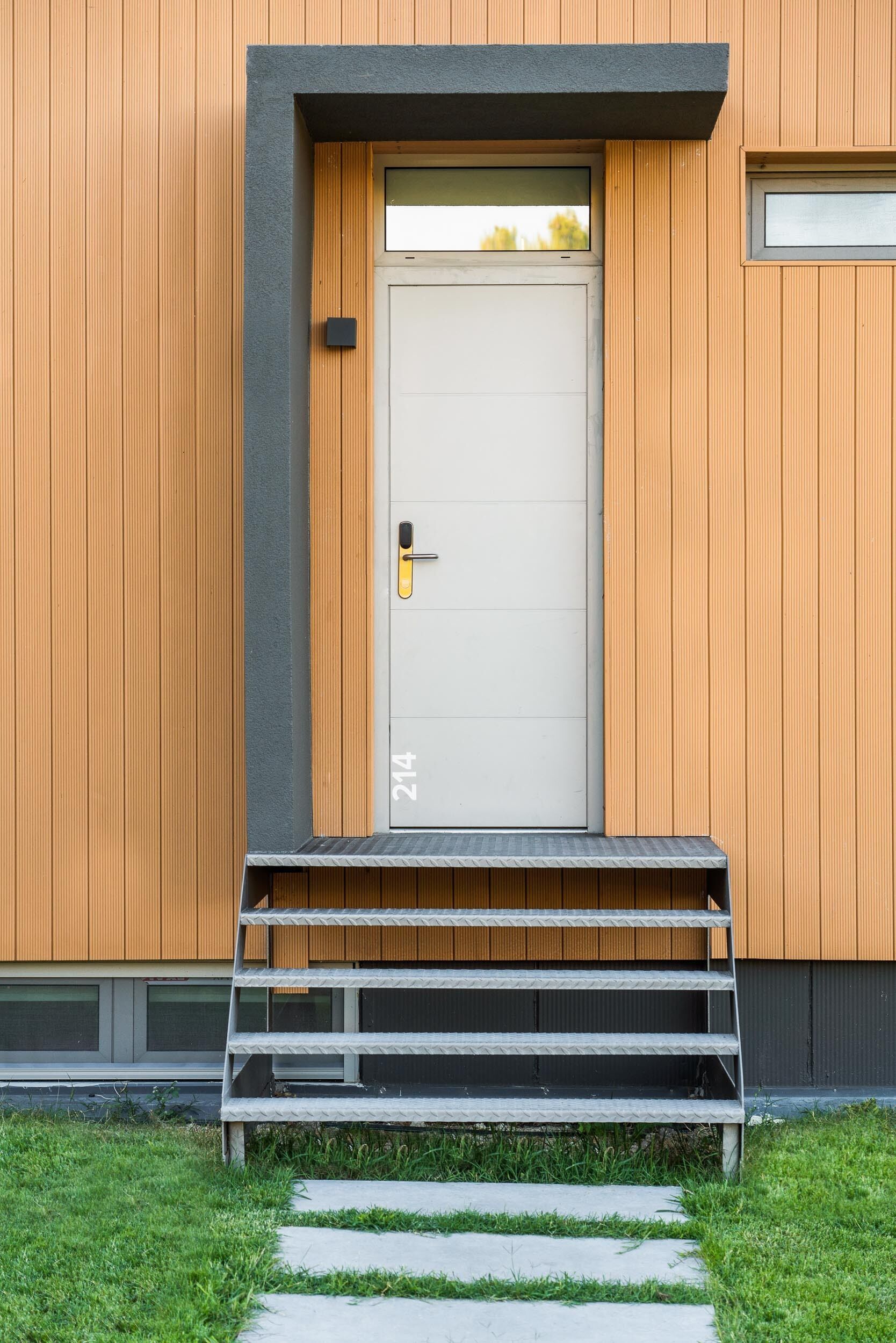 A modern building with a wooden facade, a white door with a number '214' below it, and a small set of steps leading to the entrance.
