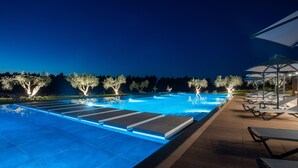 Une piscine nocturne aux bords illuminés, entourée de chaises longues et de parasols, avec des arbres décorés de guirlandes lumineuses en arrière-plan.