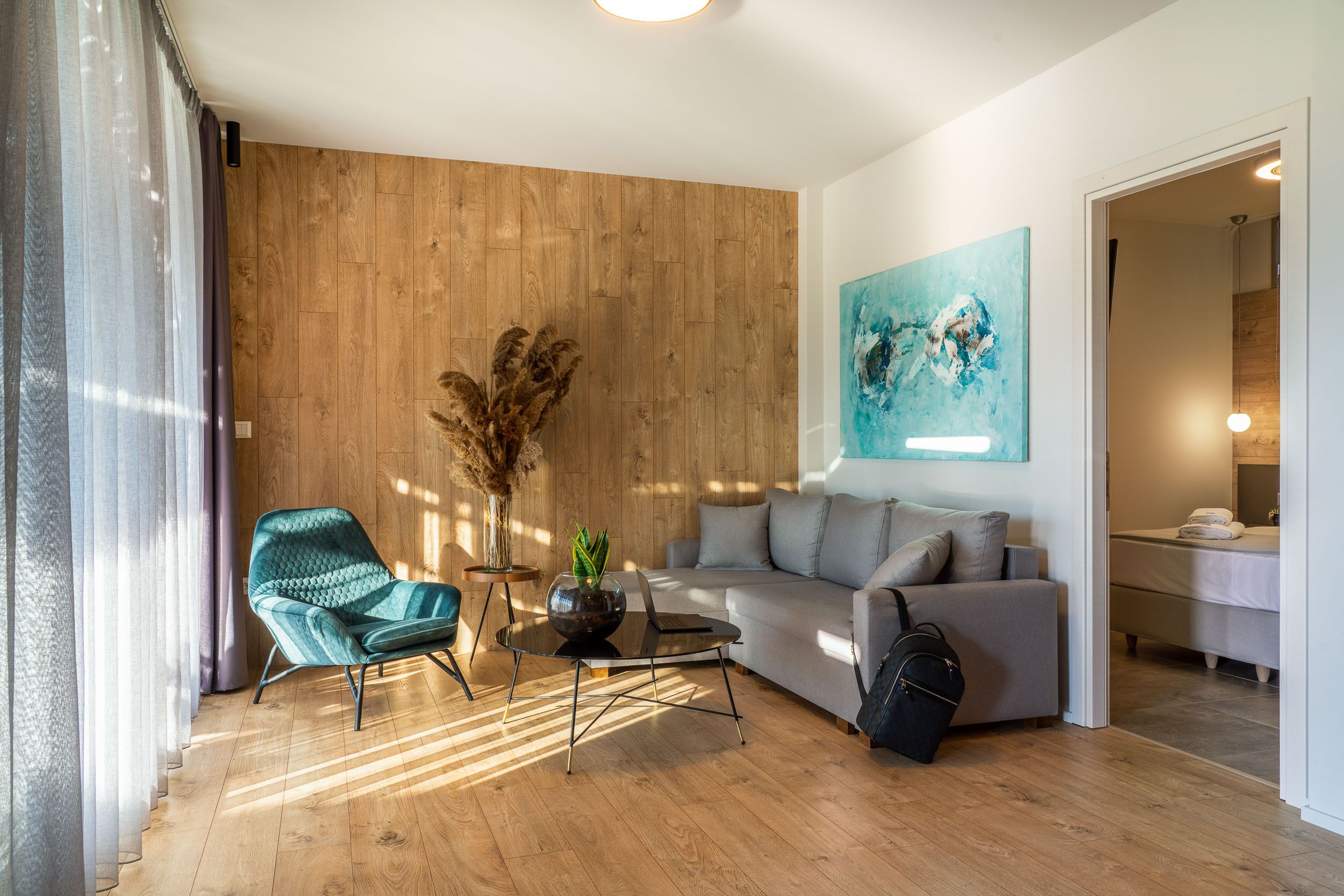 A modern living room with a wooden wall, a grey sofa, a teal armchair, a glass coffee table, and a painting.