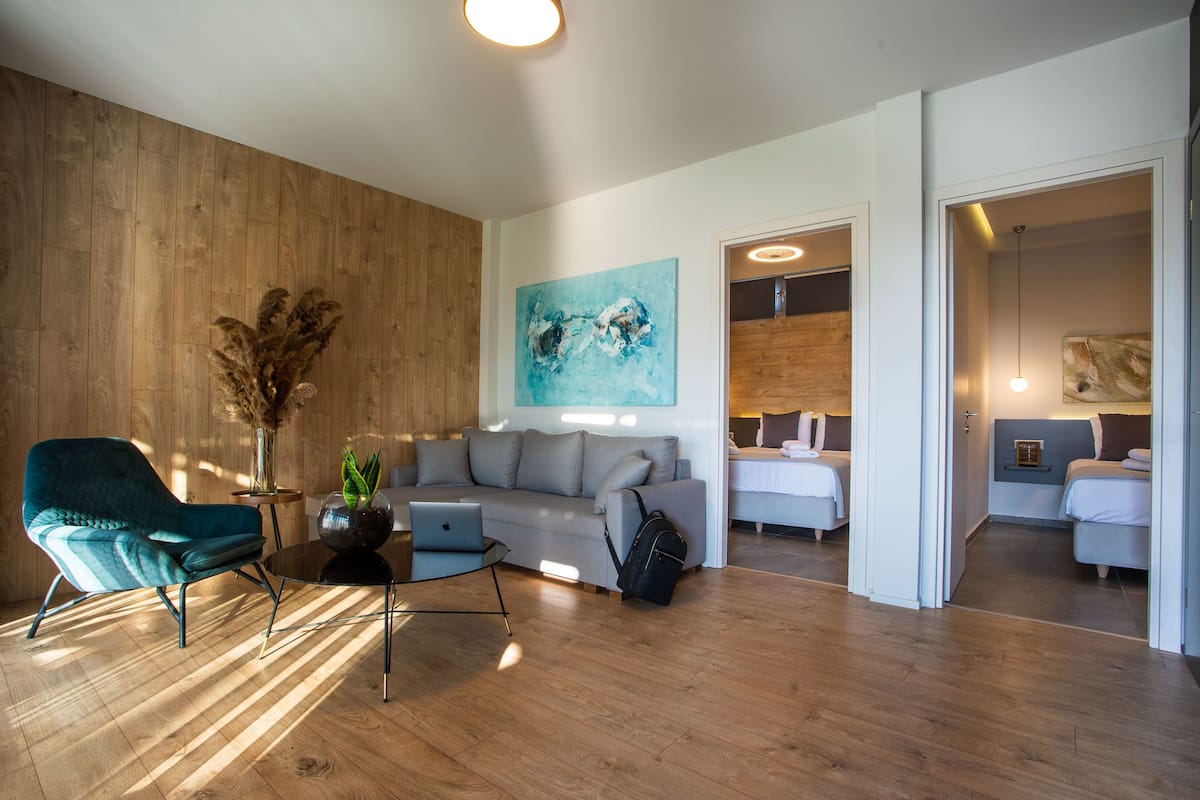 Ein modernes Wohnzimmer mit einer Couch, einem Türkis-farbenen Sessel und einem Couchtisch. An der Wand hängt ein Gemälde, und eine Tür führt in ein Schlafzimmer.