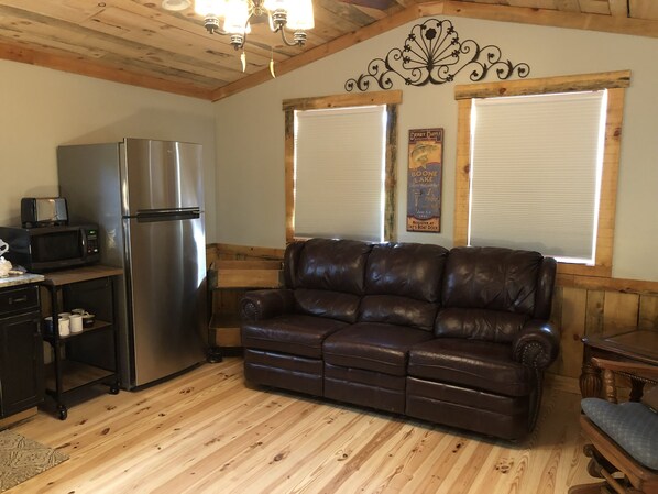 A cozy cabin interior with a kitchenette, a leather sofa, wooden floors, and a decorative wall piece.