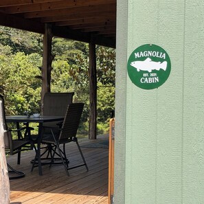 A wooden deck with a table and chairs, a green sign with a fish silhouette, and a forested background. - Magnolia Cabin (Johnson City)