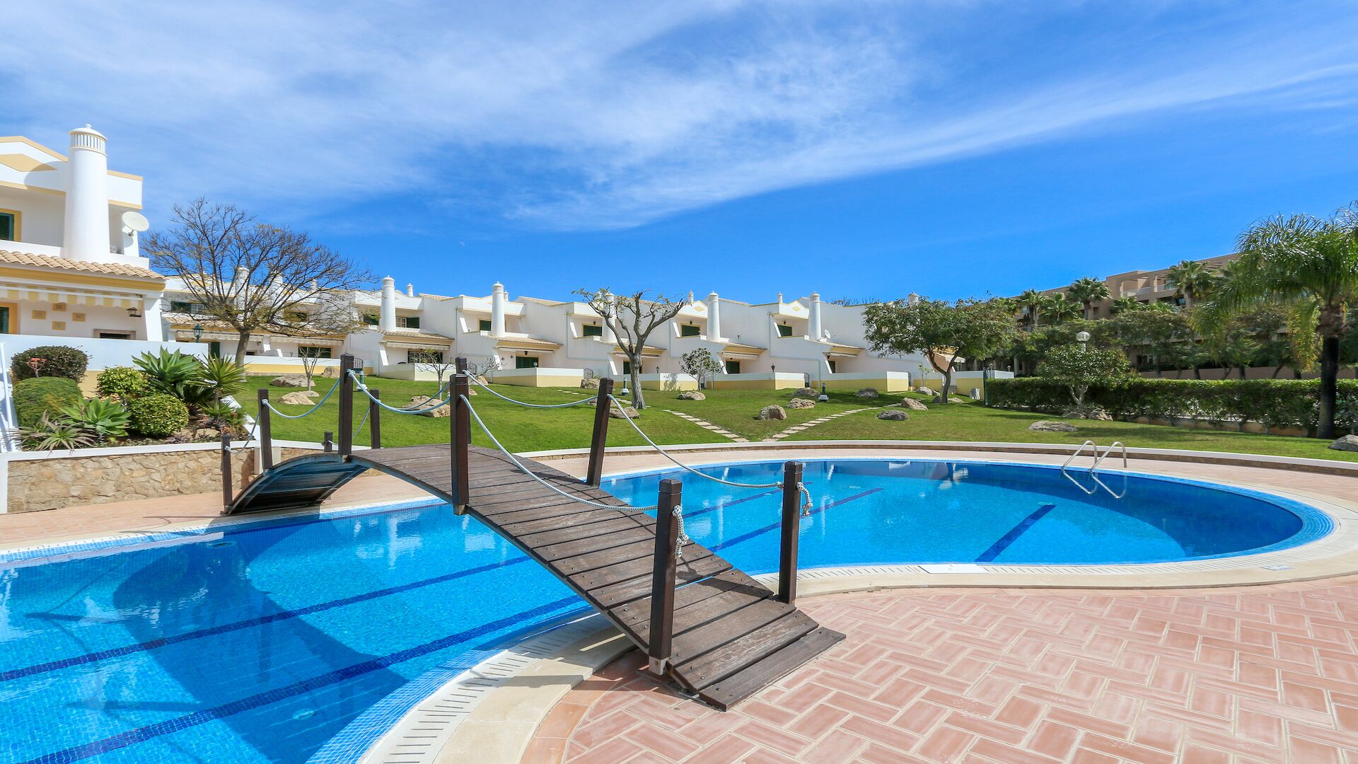 Una piscina con cubierta de madera, rodeada por un césped cuidado y una hilera de edificios blancos con balcones.