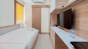 A compact hotel room with a bed, a TV, a desk with a remote control, and a water bottle. - Comfort and Cozy Studio Apartment at B Residence (Pagedangan)