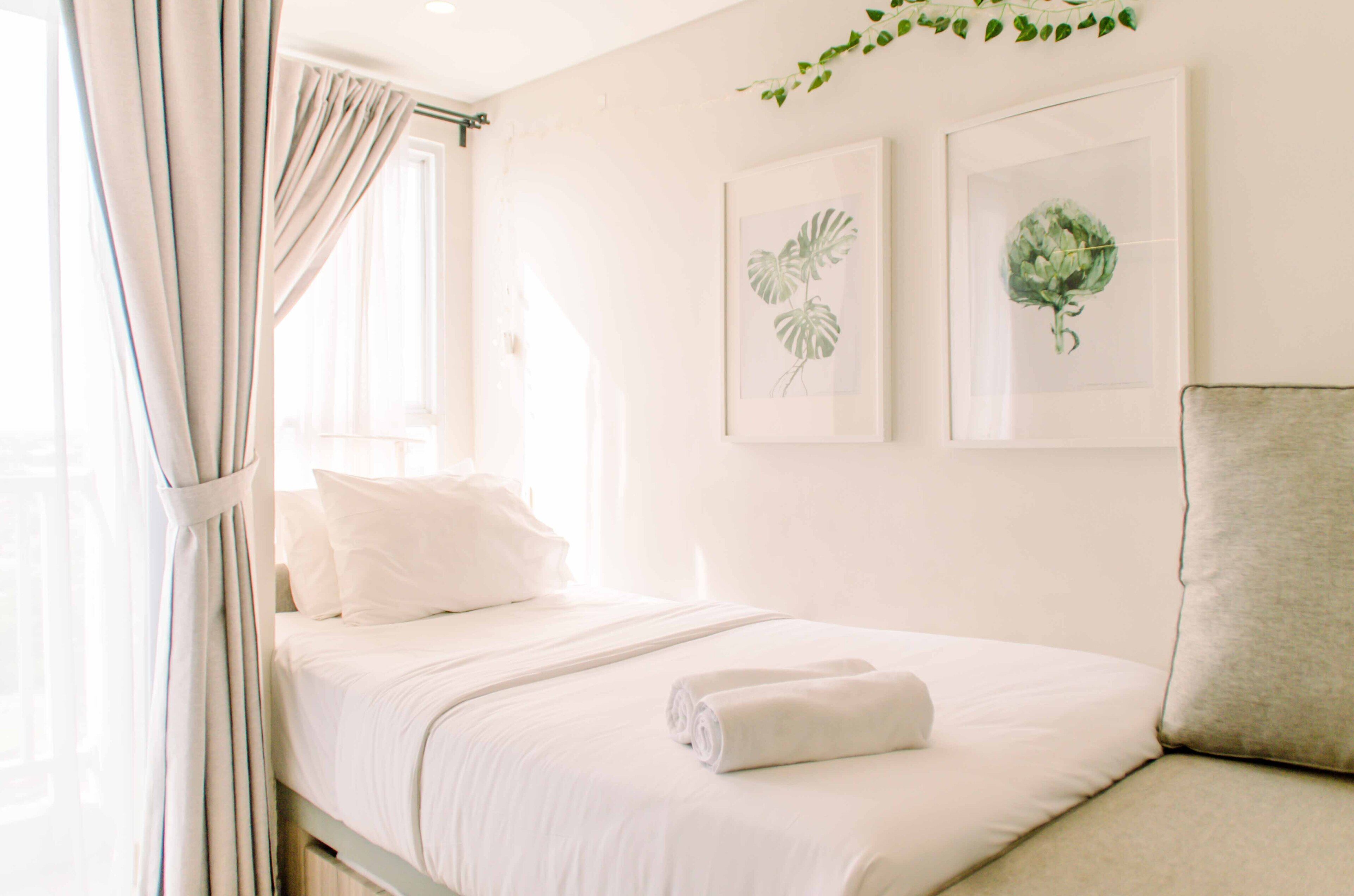 A neatly made bed with white linens and a rolled towel, a window with sheer curtains, and two framed botanical prints on the wall.